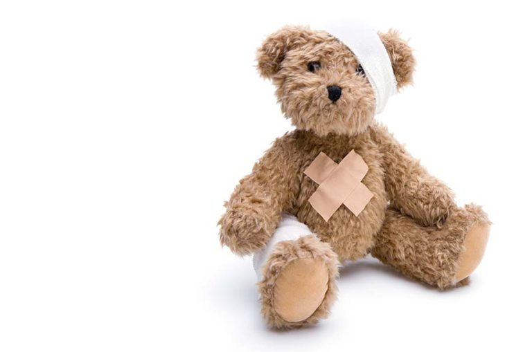 A teddybear with a head and belly bandage sits on against a white background.
