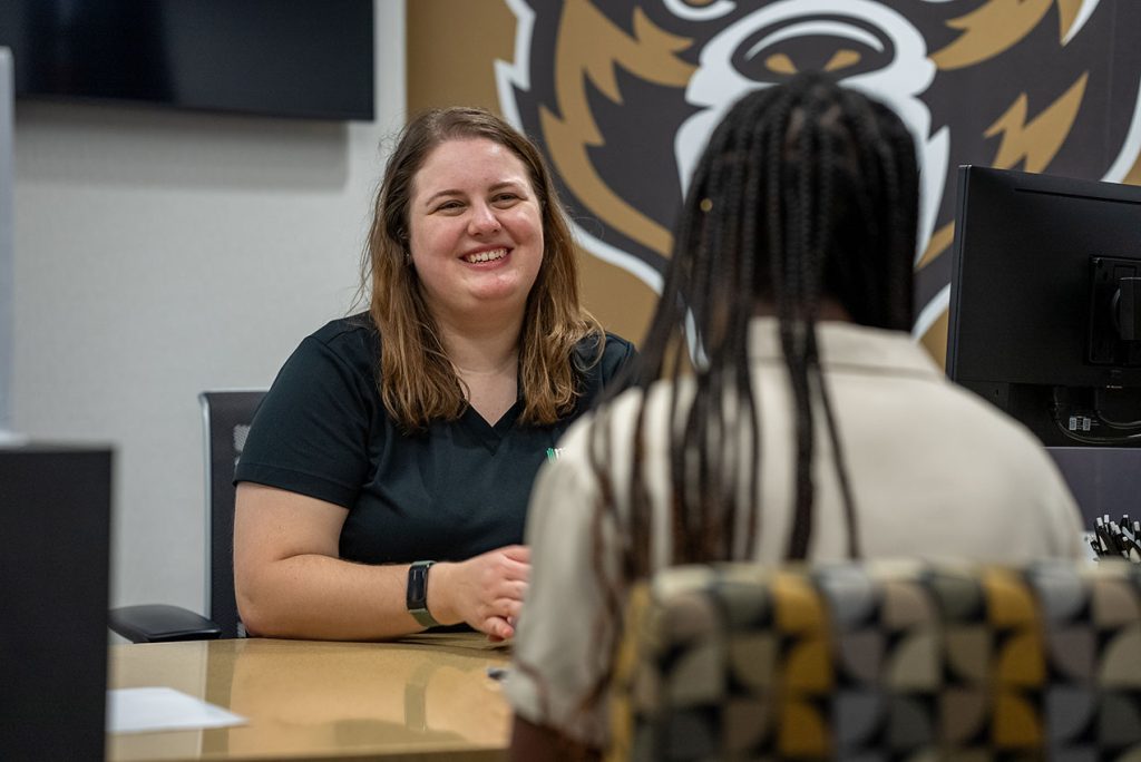 A Credit Union employee with long brown hair smiles while talking to an individual with long black hair braids.