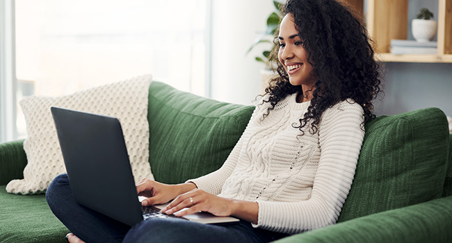Smiling person with long dark hair sits crosslegged on a green couch while using a laptop.